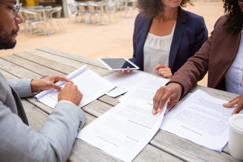 Three professionals sit at an outdoor table reviewing documents, including business contracts and proposals, during a discussion—symbolizing the importance of background checks in responsible hiring decisions.